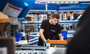 Ein junger Mann in einem AUTODOC-T-Shirt verpackt ein AUTODOC-Paket in einem Lager.