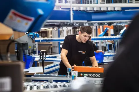 Ein junger Mann in einem AUTODOC-T-Shirt verpackt ein AUTODOC-Paket in einem Lager.