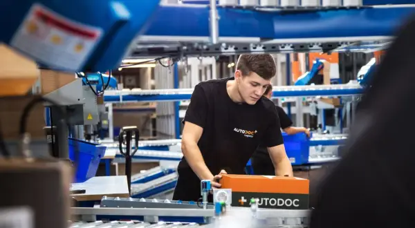 Ein junger Mann in einem AUTODOC-T-Shirt verpackt ein AUTODOC-Paket in einem Lager.